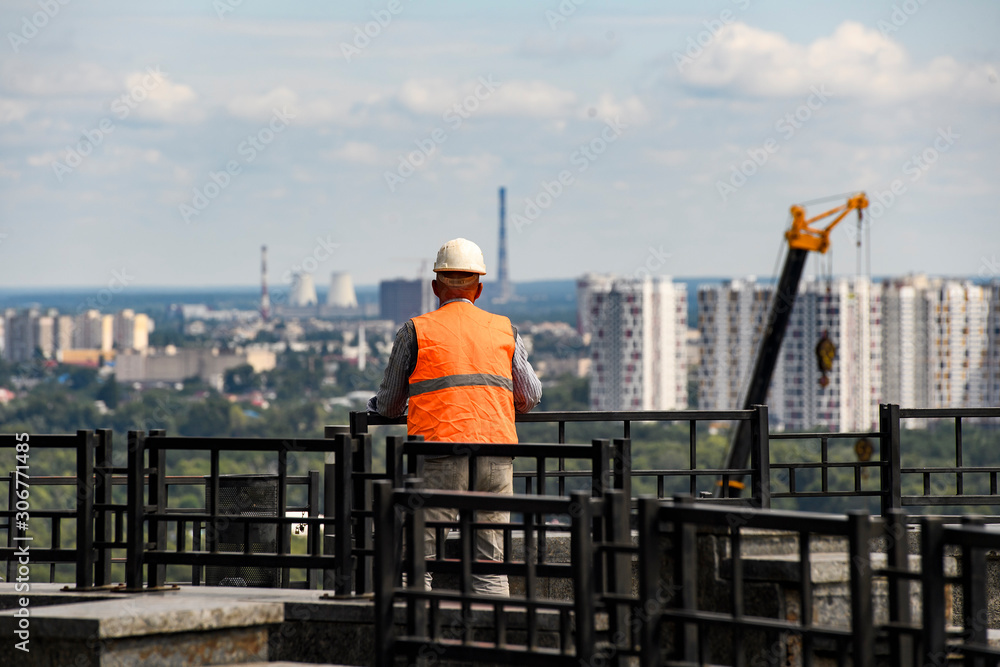 Builder worker in a helmet looks at the city with cranes and houses.