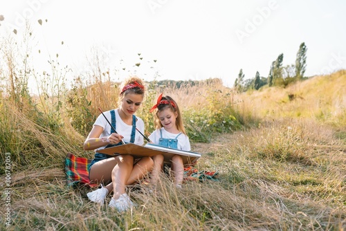 Beauty mother paint with her little daughter. Stylish woman drawing the picture with little girl. Cute kid in a white t-shirt and blue jeans. Mother's Day