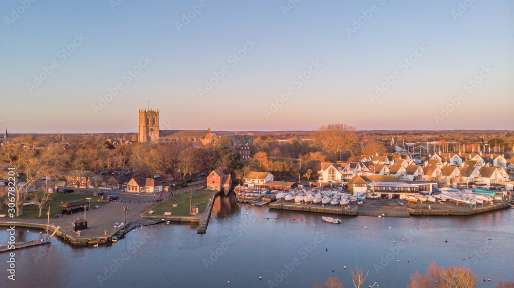 An aerial view of a river at golden hour with grassy bank and majestic church and marina in the background under a majestic blue sky
