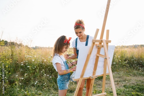 Beauty mother paint with her little daughter. Stylish woman drawing the picture with little girl. Cute kid in a white t-shirt and blue jeans. Mother's Day