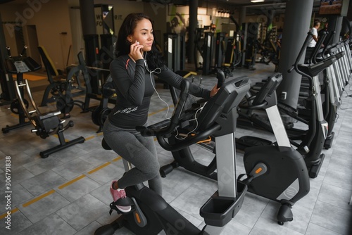 Young woman with headphones doing exercises on stationery bicycle in a gym or fitness center. Young sporty woman in gym listen music from smartphone. Women doing cardio exercises.
