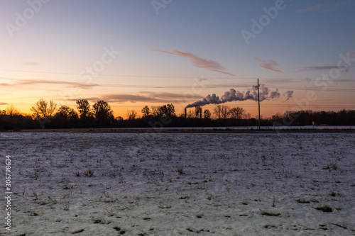 Sunrise over a snow covered field with a factory smokestack blowing in the distance