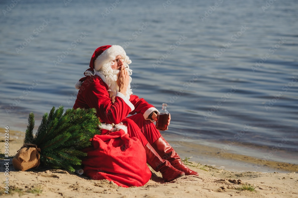 Obraz premium Santa Claus on the beach drinks whiskey and smokes a cigar.