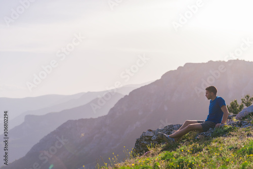 Adventurous man sitting on top of a mountain and enjoying the beautiful view, while looking downhill at the blue river and amazing mountain line..