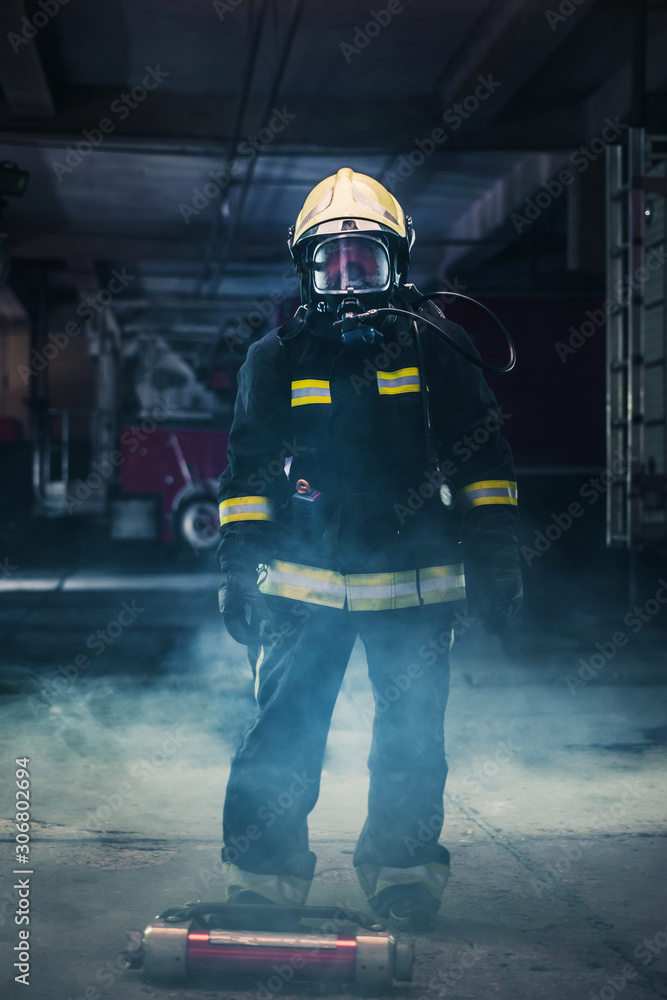Fototapeta premium Portrait of a female firefighter wearing a helmet and all safety equipment a while holding a tomahawk and wearing an oxygen mask indoors surrounded by smoke