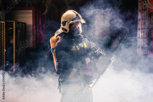 Portrait of young fireman standing and holding a chainsaw in the middle of the chainsaw's smoke .