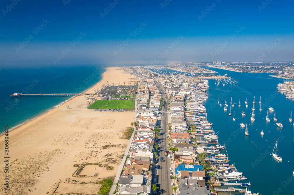 Aerial drone shot over Newport Beach in Orange County, California with pier, beach, sand and coastal neighborhood and homes on a sunny blue sky day.