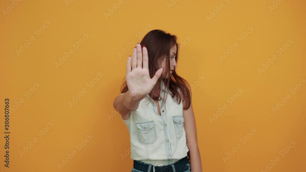 Attractive brunette caucasian young woman in blue denim shirt doing stop gesture isolated on orange background in studio. People sincere emotions, lifestyle concept. Attractive brunette caucasian young woman in blue denim shirt doing stop gesture isolated on orange background in studio. People sincere emotions, lifestyle concept.
