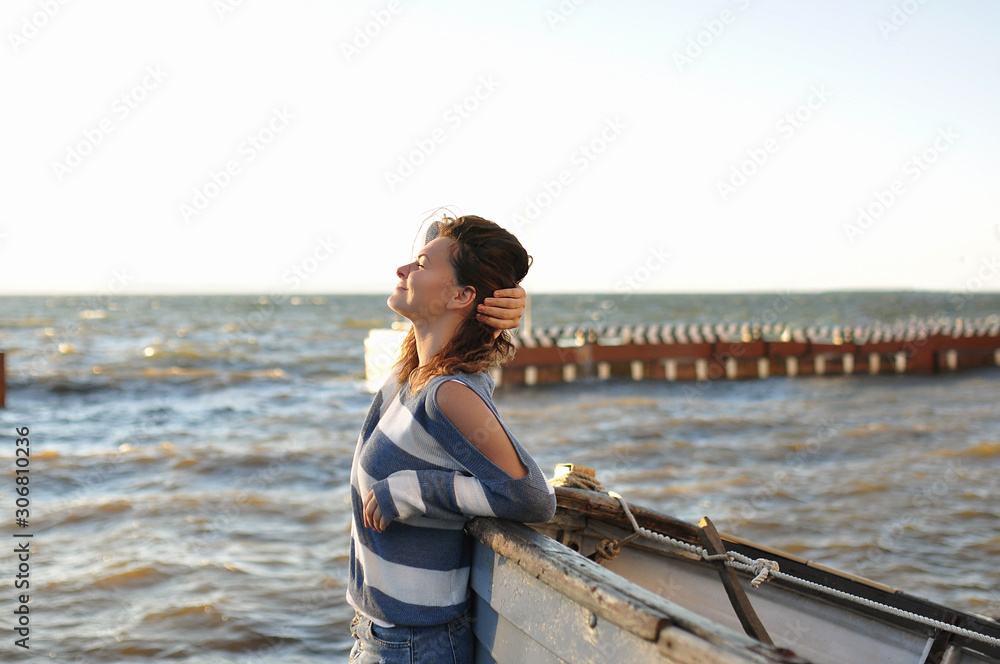 brunette girl in a striped sweater, bare shoulder and a denim skirt with holes, smiling near an old gray boat on a background of the sea, enjoying nature