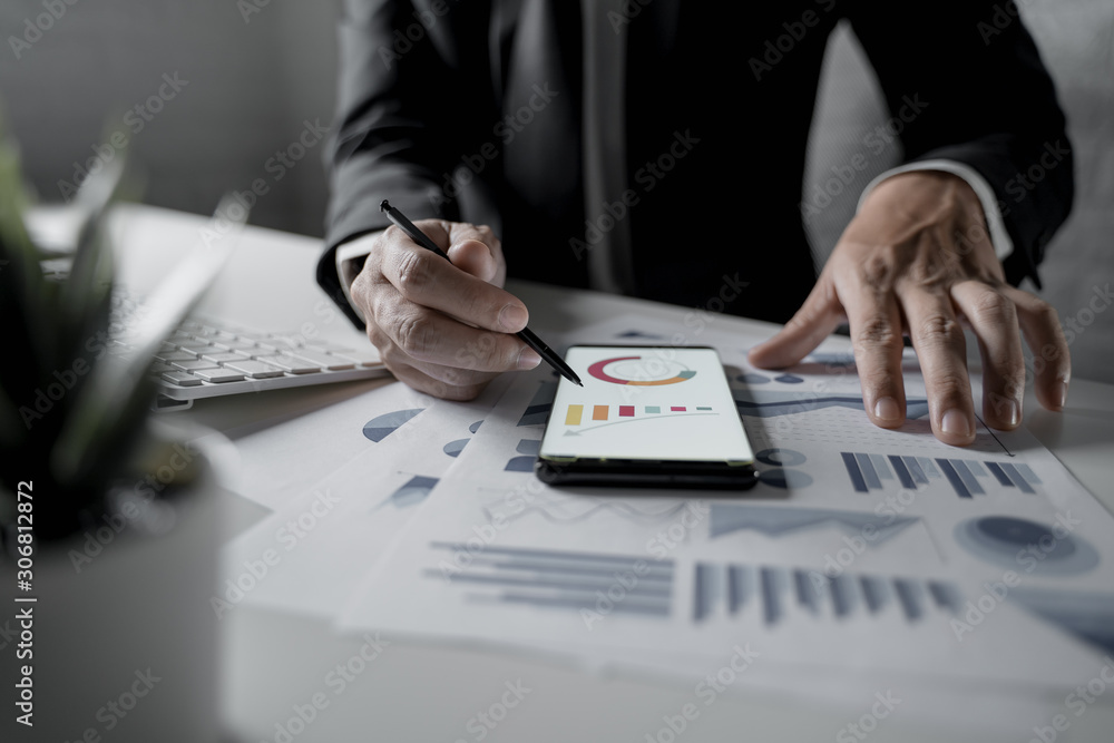 Close up shot of businessman working in office holding smart phone and checking diagrams Close up shot of businessman working in office holding smart phone and checking diagrams