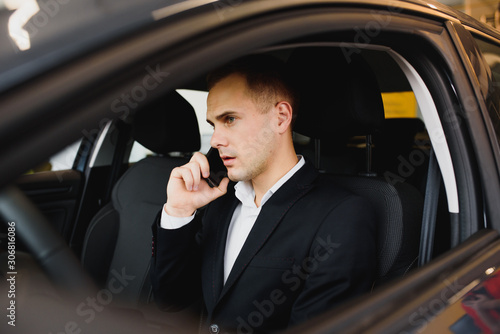 Young businessman sits in luxury car and talks on phone. He looks straight forward. Guy drives car. He holds one hand on steering wheel. It is sunny outside. © Serhii