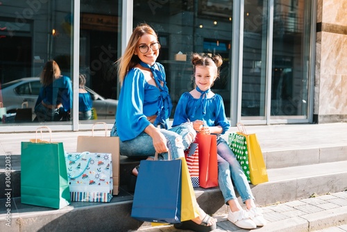 Beautiful mom and her cute little daughter are holding shopping bags, looking at camera and smiling while standing outdoors. Shopping concept.