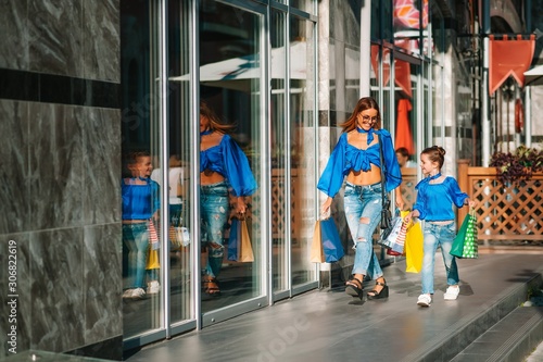 Beautiful mom and her cute little daughter are holding shopping bags, looking at camera and smiling while standing outdoors. Shopping concept.