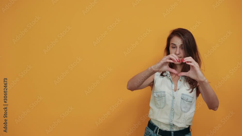 Attractive caucasian young woman in blue denim shirt showing shape of heart gesture isolated on orange background in studio. People sincere emotions, lifestyle concept. Attractive caucasian young woman in blue denim shirt showing shape of heart gesture isolated on orange background in studio. People sincere emotions, lifestyle concept.