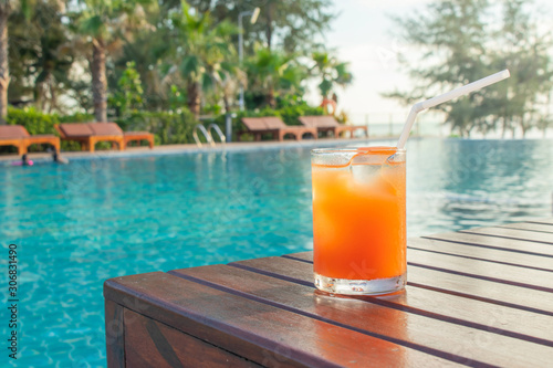 Summer Vacation Concept : Welcome drink orange juice punch put on wooden table nearly swimming pool with blue water and daybed in background.