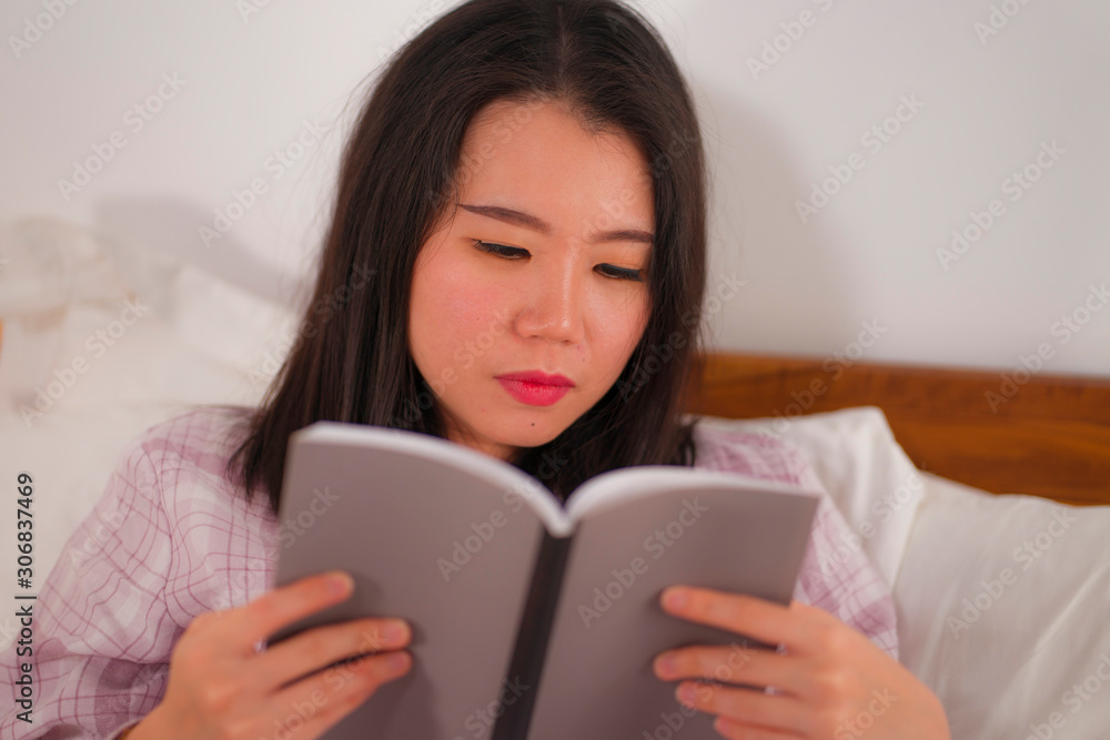 young beautiful and sweet Asian Korean woman in bed reading book novel feeling happy and relaxed or studying at home wearing cute pajamas cozy and homey