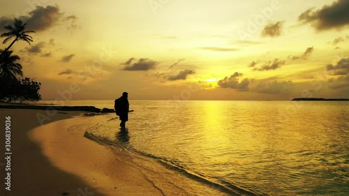 Golden sunset with yellow sun setting down the grey clouds, reflecting over calm glassy surface of sea washing pristine beach with bent palms, silhouette of girl 