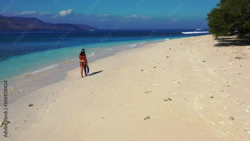 Girl in swimsuit with snorkel set walking on quiet exotic beach alongside waves of blue turquoise sea, searching for right place to dive