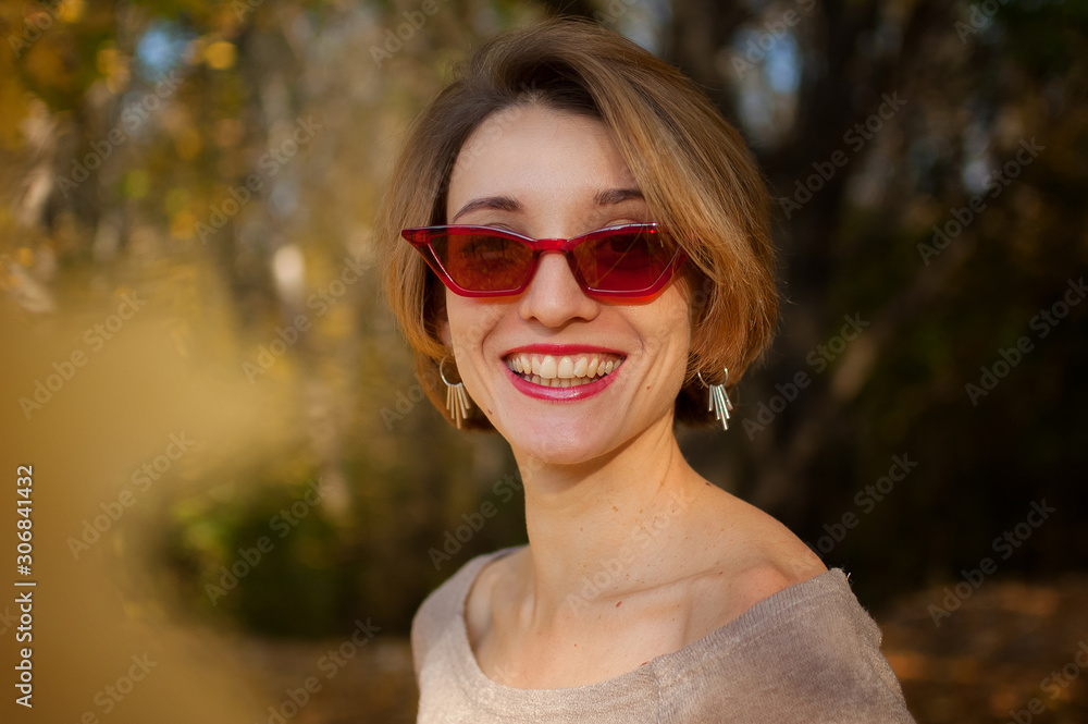 Young and beautiful girl with short hair and red fashionable sunglasses is posing against the trees with yellow leaves background spending time in the autumn park Young and beautiful girl with short hair and red fashionable sunglasses is posing against the trees with yellow leaves background spending time in the autumn park