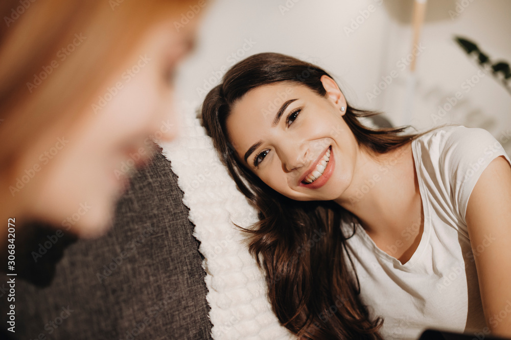 Fototapeta premium Portrait of a lovely young woman with long dark hair laughing while leaning head on the sofa looking at her female friend with red hair.