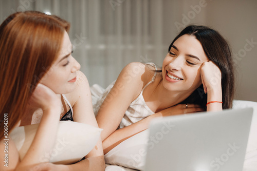 Two beautiful young girlfriend having fun while leaning on a bed with a laptop in the front of their face looking to each other laughing. photo