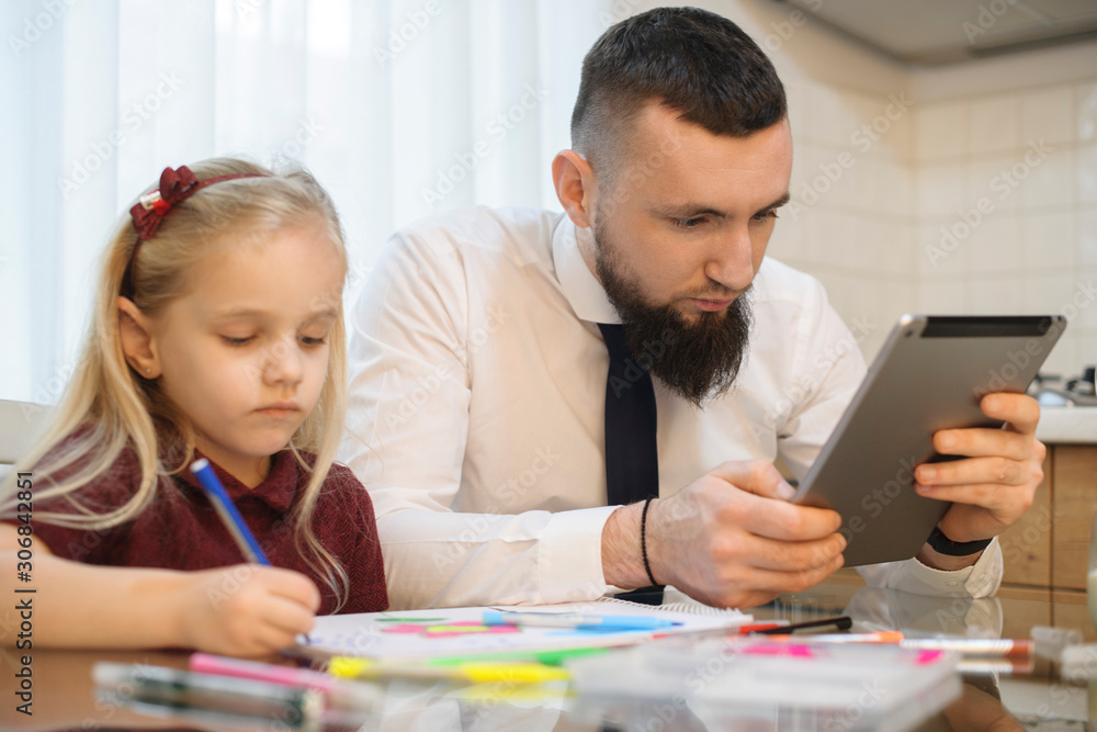 Father looking at a tablet screen in the kitchen before going to work while his little girl si drawing. Father looking at a tablet screen in the kitchen before going to work while his little girl si drawing.
