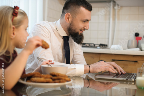 Side view portrait of a young bearded father dressed in suit before going to work looking at his laptop in the kitchen while his daughter is eating biscuits.