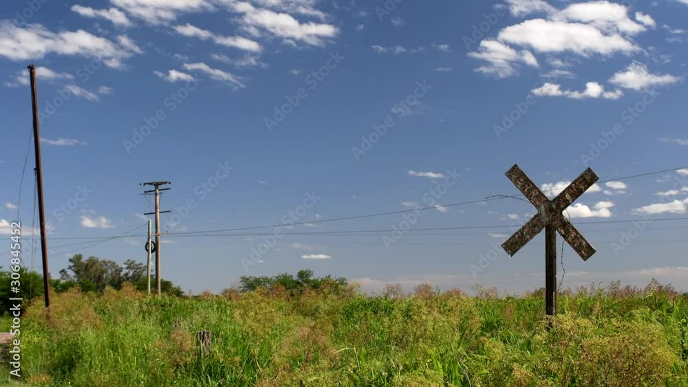 A view featuring an old railroad warning on an abandoned track, poles and cables and a clear blue sky spotted with some clouds, A view featuring an old railroad warning on an abandoned track, poles and cables and a clear blue sky spotted with some clouds,