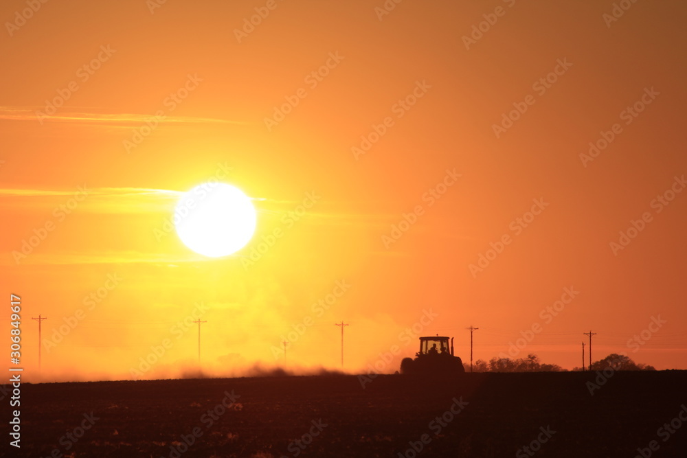 Fototapeta premium Kansas colorful Sunset with a farmer woking in the field stirring up dust out in the country.