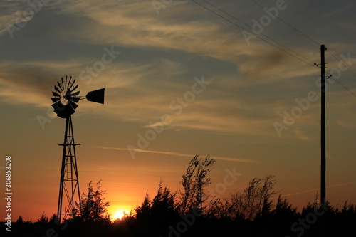windmill at sunset with clouds and tree's and the Sun with powerlines and poles.
