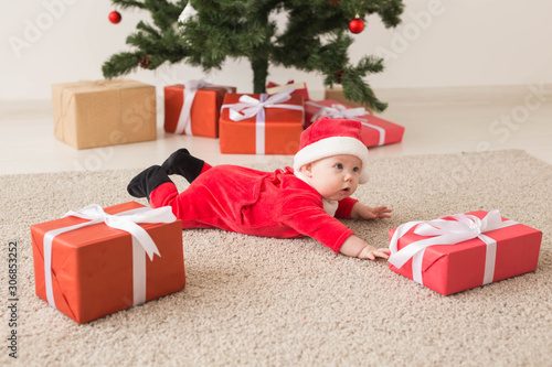 Cute baby girl wearing santa claus suit crawling on floor over Christmas tree. Holiday season.