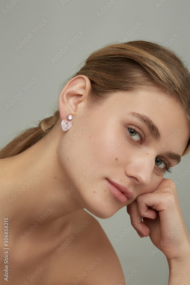Cropped close-up shot of a charming woman with a stud earring made as a sparkling crystal in a silver chase with a pinkish heart-shaped gem pendant. The lady's blonde hair is gathered in a knot. Cropped close-up shot of a charming woman with a stud earring made as a sparkling crystal in a silver chase with a pinkish heart-shaped gem pendant. The lady's blonde hair is gathered in a knot.