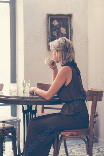 Toned picture of side view of beautiful blonde lady sitting in cafe or restaurant near window and drinking coffee