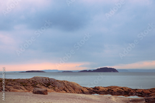 Obraz na płótnie Landscape of Oslo Fjord with small islands, calm water, overcast sky. orange rocks on the beach, tranquility. Pastel colors. Norwegian coastline in summer. Nesodden Norway. Nesoddtangen peninsula. w salonie