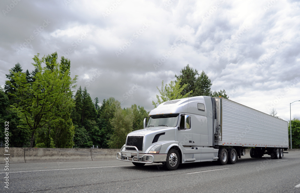 Bonnet big rig semi truck with grille guard transporting cargo in refrigerator semi truck running on the wide highway with trees on the background Bonnet big rig semi truck with grille guard transporting cargo in refrigerator semi truck running on the wide highway with trees on the background