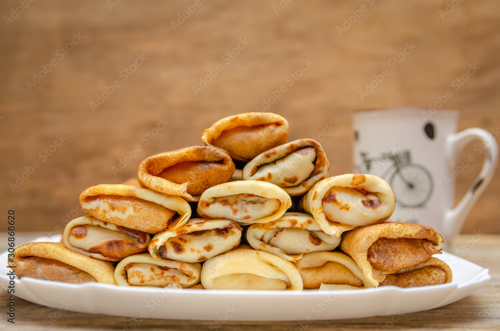 Pancakes with filling are a pyramid on a white plate standing on a wooden background close-up, in the background a Cup