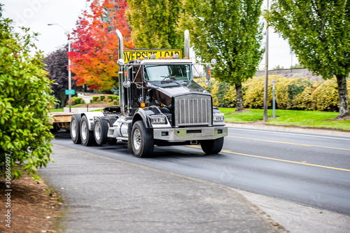 Powerful big rig black semi truck with step down semi trailer and sign for transporting oversize load running on the local road with autumn trees on the side