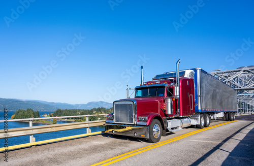Red classic bonnet big rig semi truck transporting frozen cargo in bright refrigerated semi trailer driving on the truss bridge across the river