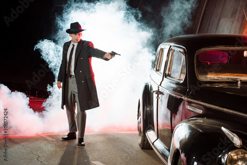 stylish gangster in hat holding gun near retro car and smoke