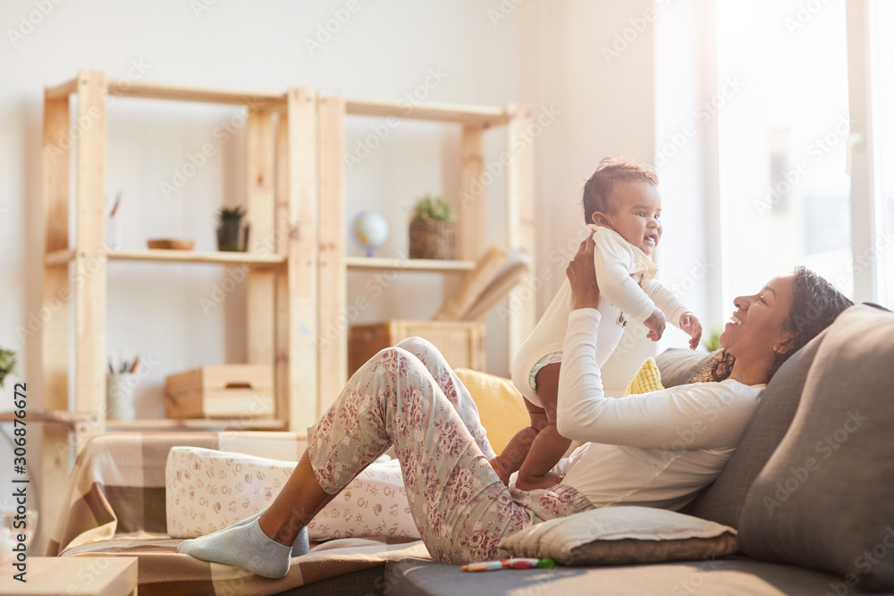 Warm toned portrait of young African-American mother playing with baby on couch lit by sunlight, copy space Warm toned portrait of young African-American mother playing with baby on couch lit by sunlight, copy space