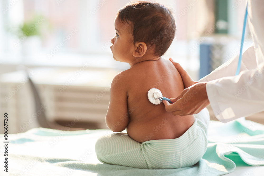Rear view of shirtless toddler sitting on the table while doctor listening to him with stethoscope - obrazy, fototapety, plakaty 