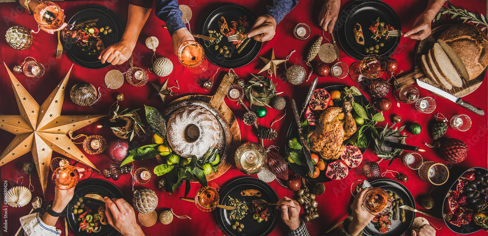 Friends celebrating Christmas. Flat-lay of people feasting over festive table with red cloth with champagne, roasted chicken, bundt cake, fruits and decorations, top view. Winter holiday party