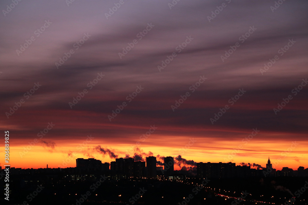 Sunrise over the city, scenic view with orange dramatic sky. Dark storm clouds and black smoke above silhouettes and lights of high-rise buildings