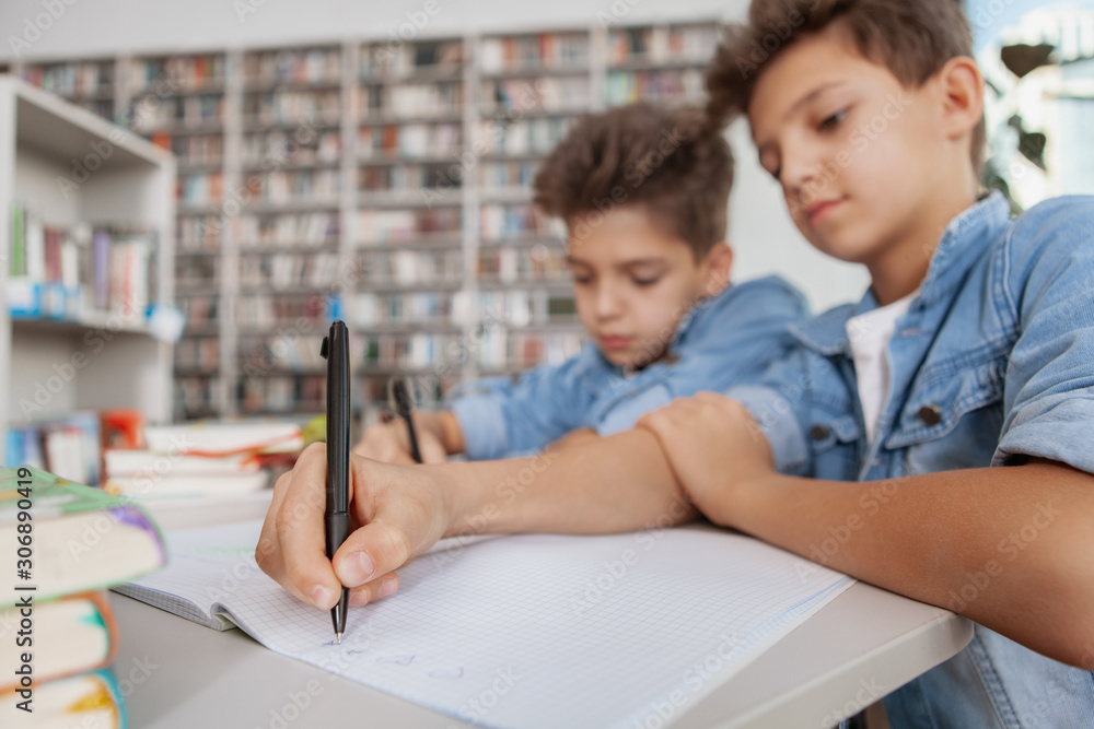 Twin brothers doing homework together. Young boys studying at the library writing in textbooks, copy space Twin brothers doing homework together. Young boys studying at the library writing in textbooks, copy space