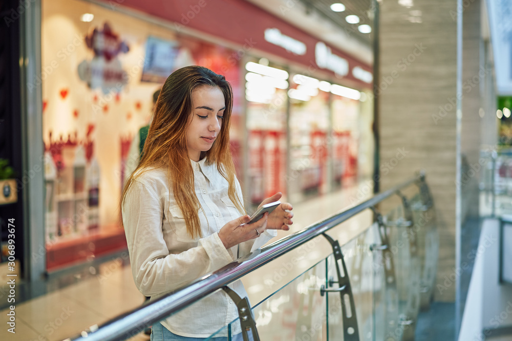 young woman in a shopping center stands near the fence and using app  on the phone