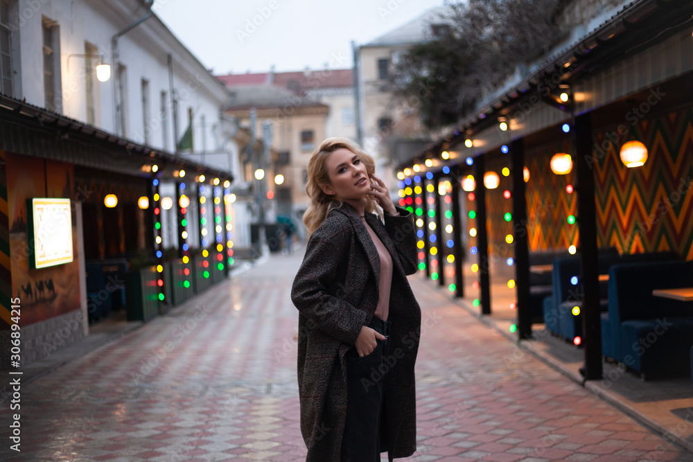 Fashion woman walking at the city, dressed modern clothes. Female model wearing coat, tutleneck, trousers and posing against the street with light at the evening