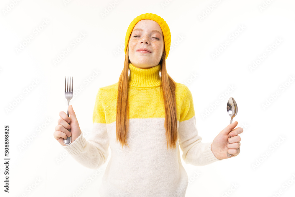surprised european red-haired girl holds a fork and knife on a white wall surprised european red-haired girl holds a fork and knife on a white wall