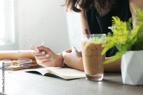 Closed up Asian woman in black dress holding pencil and thinking on blank notebook with eyeglasses, Iced coffee and old books on dark working table with warm light window.