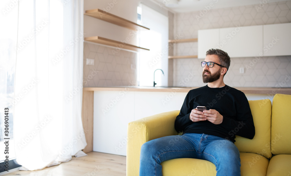 Happy mature man sitting on sofa in unfurnished house, holding smartphone.