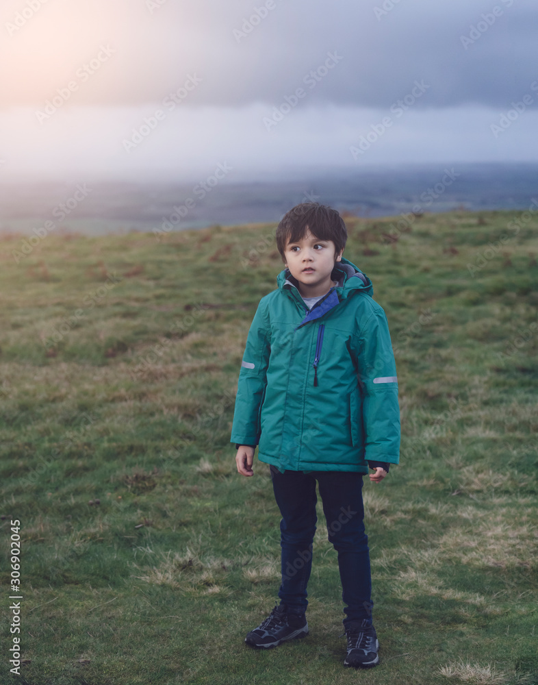 Portrait of little boy standing alone and looking out with thingking face with blurry top of the hill background, 4-5 years old kid wearing warm cloths and blue jean posing on the field in winter.